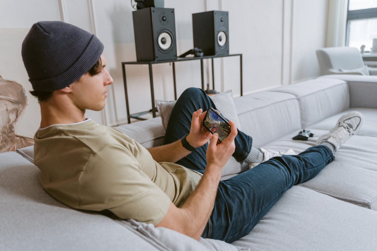 Casual young man playing mobile games on sofa with speakers in the background.